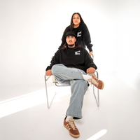 Two people wearing Te Kara – Toitū te Tiriti crewneck with visible branding in a minimalistic setting.