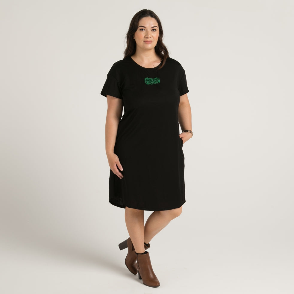 Women's black dress with stunning embroidered green maori tiki, dress has pockets and drop hem front and rear. Made in Aotearoa Tauranga New Zealand.  